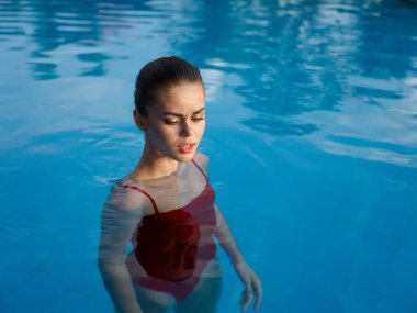 Kırmızı mayo giymiş genç bir kadın şeffaf havuz suyunun içinde çıplak omuzlarıyla dikiliyor.