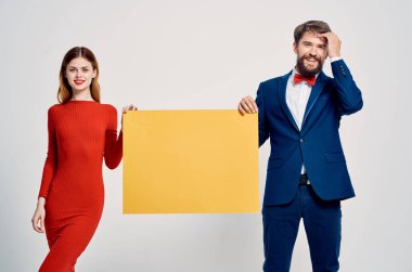 Takım elbiseli bir iş adamı kırmızı elbiseli bir kadının yanında sarı model posterler asıyor.