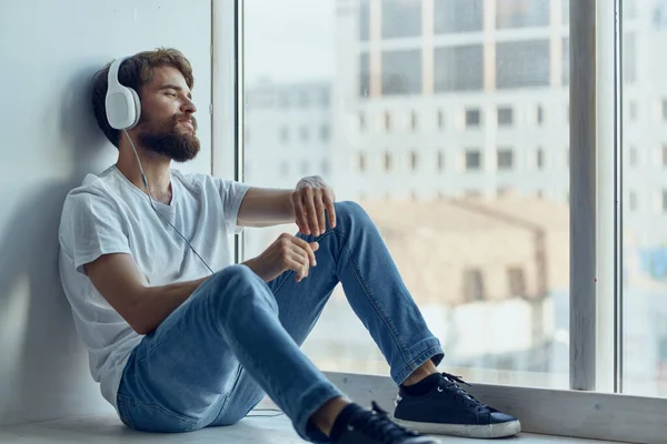 白いTシャツを着た男がヘッドフォンで窓の近くに座って音楽を聴き — ストック写真
