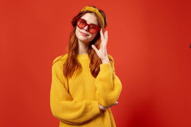 Sarı kazaklı, kırmızı gözlüklü, saç bandı takan kırmızı arka planlı bir kadın.