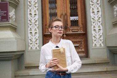 Kısa saçlı, gözlüklü bir kadın elinde bir kitapla şehirde dolanıyor.