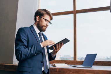 Pencerenin yanında tablet teknolojisi olan takım elbiseli bir adam var.