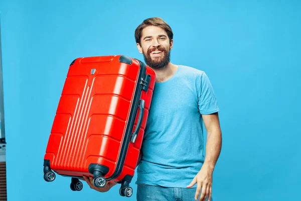 Glücklicher Mann mit Bart mit rotem Koffer in der Hand auf blauem Hintergrund — Stockfoto