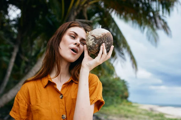 A woman in a sundress holds a coconut in her hand and green leaves of a palm tree clouds sky — Stock Photo, Image