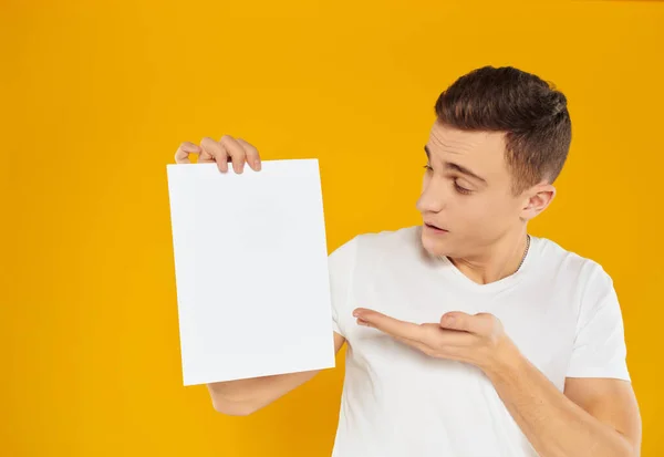 Een man in een wit t-shirt vel papier reclame kopiëren ruimte gele achtergrond — Stockfoto