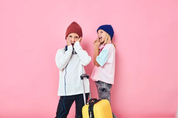 Веселый мальчик и девочка с желтым чемоданом в руках розовый цвет фона — стоковое фото