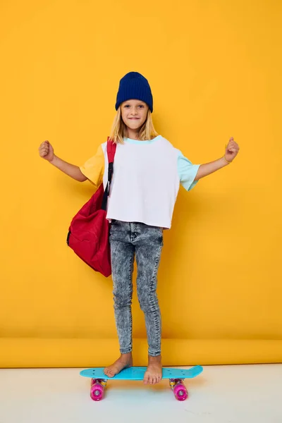 女の子の肖像画は赤いバックパックとスケートボードに乗る黄色の背景 — ストック写真