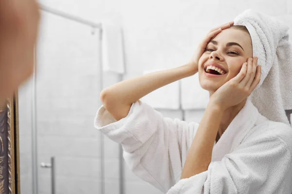 A woman looks at herself in the mirror and a towel on her head is a white robe — Stock Photo, Image