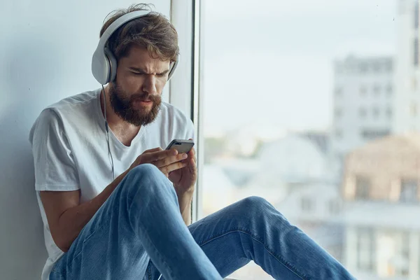 Homme en t-shirt blanc assis près de la fenêtre dans les écouteurs écouter de la musique — Photo