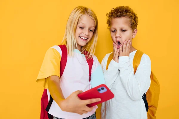 Zdjęcie dwójki małych dzieci dziewczynka z plecakami szkolnymi telefon rozrywka izolowane tło — Zdjęcie stockowe