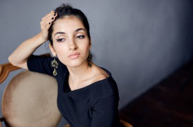 Güzel bir kadın, sandalyenin yanında duran takıları takıyor. Siyah elbise, arka plan.