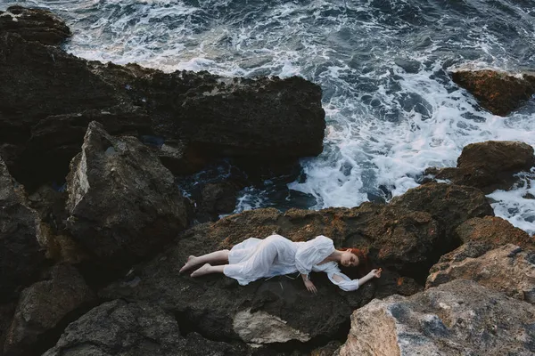 Beautiful young woman in long white dress wet hair lying on a rocky cliff landscape — Stock Photo, Image