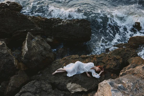 Beautiful young woman in long white dress wet hair lying on a rocky cliff landscape — Stock Photo, Image