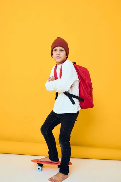 Menino com uma mochila vermelha montando um skate fundo amarelo — Fotografia de Stock