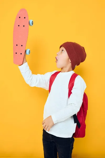 Fröhliche kleine Kind in einem roten Hut Skateboard in seinen Händen gelbe Farbe Hintergrund — Stockfoto