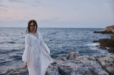 Beyaz elbiseli bir kadın okyanus kıyısındaki taşların üzerinde duruyor.