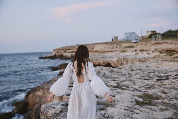 Γυναίκα με λευκό φόρεμα περπατά στην παραλία πέτρες πίσω όψη — Φωτογραφία Αρχείου