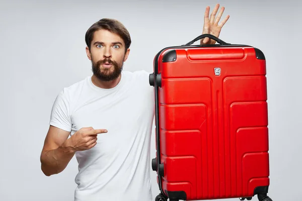 A man shows a finger to the side and a red suitcase in his hand on a light background — Stock Photo, Image