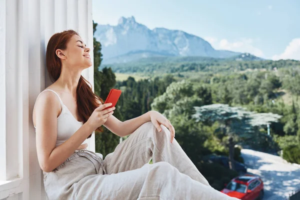 迷人的年轻女子坐在阳台上，一边打电话一边欣赏美丽的山景- -夏山景 — 图库照片
