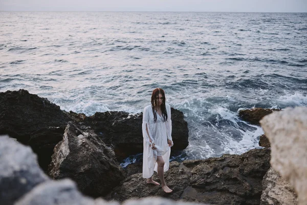 Beyaz elbiseli bir kadın okyanusun kıyısındaki kayalık taşların üzerinde duruyor. — Stok fotoğraf