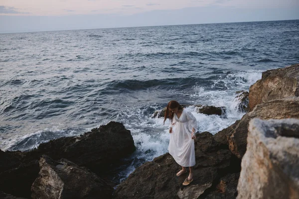 Woman climbs a stone cliff by the ocean in a white dress — Stock Photo, Image