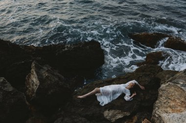 Çıplak ayaklı bir kadın, ıssız bir yerde, kayalık bir sahilde, yukarıdan beyaz bir elbiseyle.