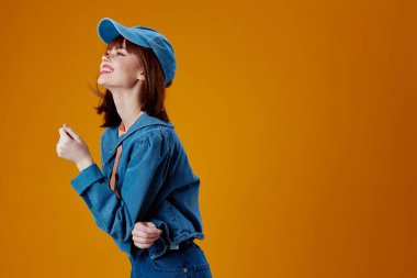 Şapka ve kot ceketli, sarı arkaplan pozu veren güzel, modaya uygun bir kız.