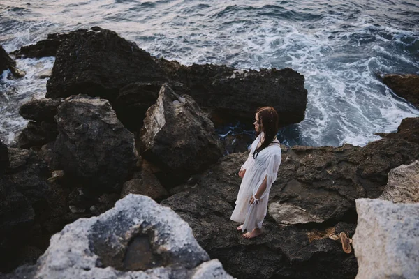 Islak saçlı beyaz elbiseli bir kadın okyanus manzaralı taşların üzerinde duruyor. — Stok fotoğraf