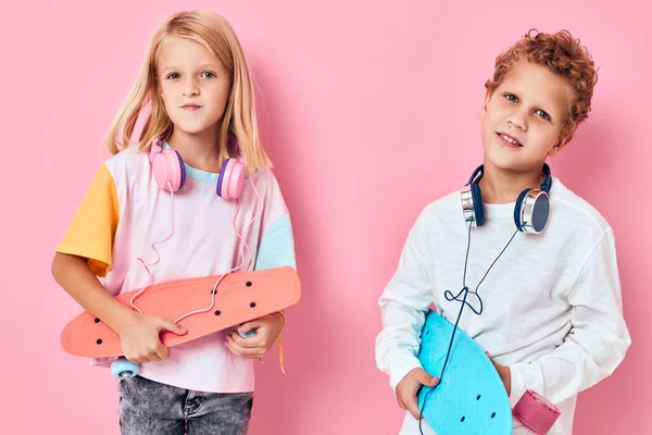 Retrato de un niño y una niña en auriculares patinetas en las manos fondo rosa —  Fotos de Stock