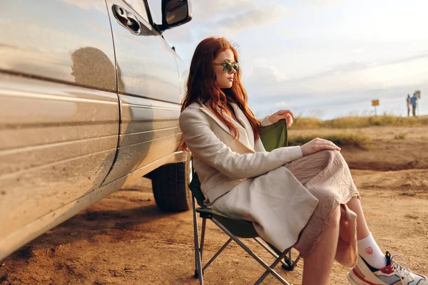 Piękna kobieta siedzi na krześle na polu w ciemnych okularach w pobliżu samochodu niekończące się pole — Zdjęcie stockowe