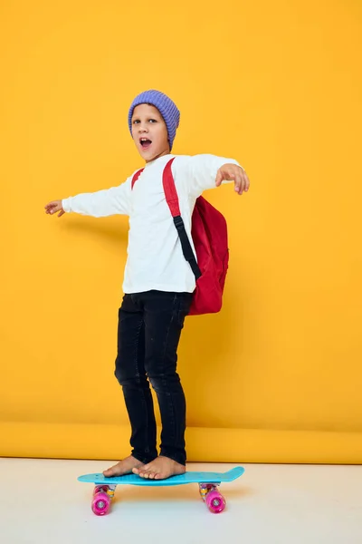 Porträt von niedlichen Jungen mit rotem Rucksack blaues Skateboard gelb Hintergrund — Stockfoto