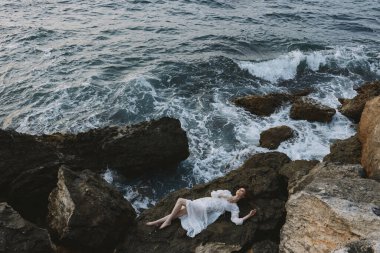 Uzun beyaz elbiseli, ıslak saçlı, değişmemiş kayalık bir uçurumda uzanan güzel bir kadın.