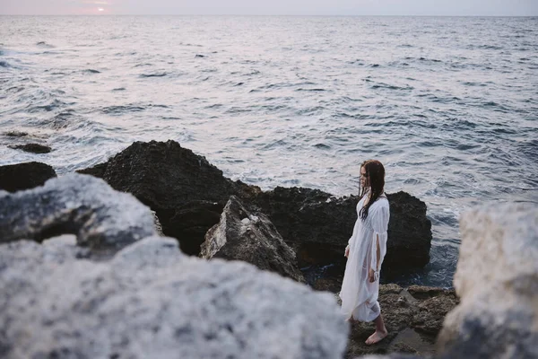 Kadın beyaz elbiseli kayaların üzerinde duruyor. Okyanus doğası değişmeden seyahat ediyor. — Stok fotoğraf