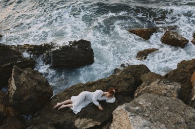 Beyaz elbiseli çıplak ayaklı kadın, beyaz elbiseli tatil konseptiyle bir taşın üzerinde yatıyor.