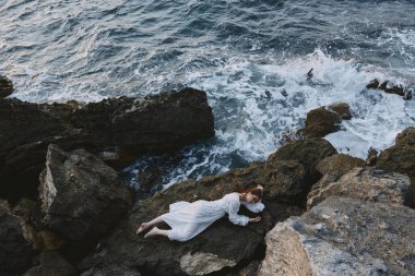 Uzun beyaz elbiseli, çıplak ayaklı, ıslak saçlı bir kadın kayalık bir uçurum kenarında yatıyor.