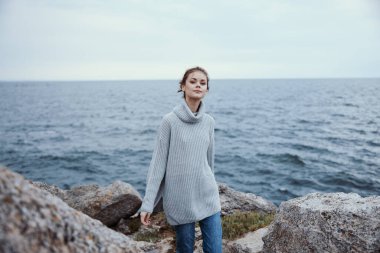 Gri kazaklı güzel bir kadın kayalık bir sahil doğasında hayat tarzında duruyor.