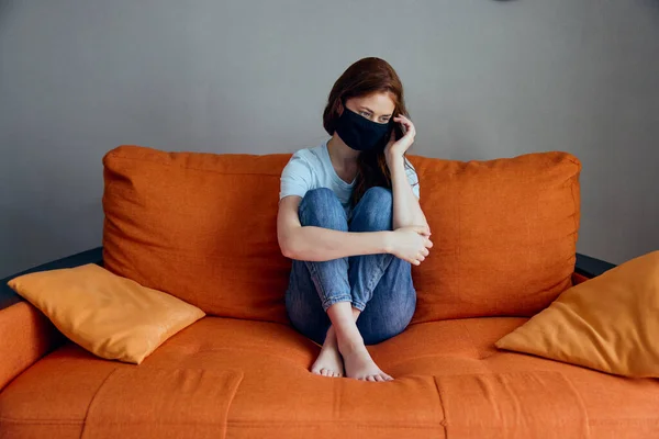 Smutná žena sedí na oranžové pohovce doma v ochranných maskách životní styl — Stock fotografie