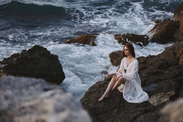 Mujer vestida de blanco se sienta en una piedra cerca de las olas del océano viajar por la naturaleza —  Fotos de Stock