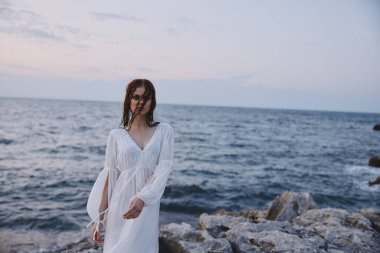 Woman in white dress and wet hair stony ocean shore