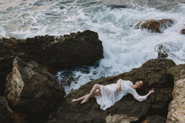 Beyaz elbiseli, uzun saçlı çekici bir kadın. Beyaz elbiseli, taşın üzerinde yatıyor.