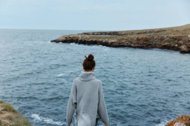 woman in a gray sweater standing  on a rocky shore 