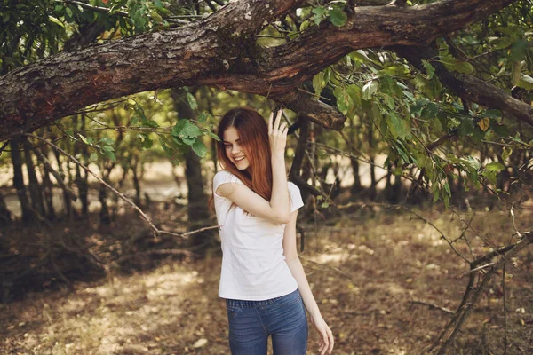 Жінка в футболці і джинсах стоїть біля дерева з зеленим листям на природі в парку — стокове фото