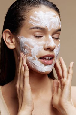 Güzel kadın cilt bakımı için yüz bej arka planda beyaz maske kullanılıyor.