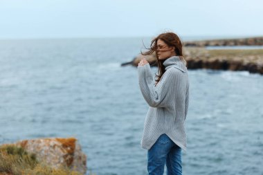 Gri kazaklı güzel bir kadın kayalık bir sahil doğasında hayat tarzında duruyor.