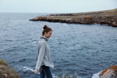 Gri kazaklı kadın kayalık bir kıyıda duruyor. Kadın dinleniyor.