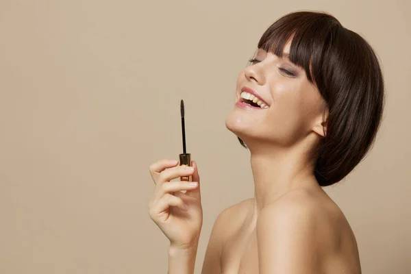 Woman paints eyelashes bare shoulders beige background — Stock Photo, Image