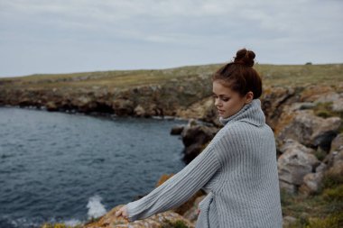 Gri kazaklı kadın kayalık bir sahil doğasında hayat tarzında duruyor.