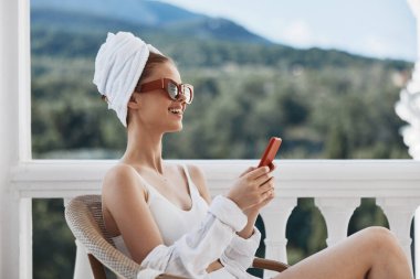 Güneş gözlüklü çekici genç kadın balkonda oturuyor ve tembel bir sabah telefonu kullanıyor.