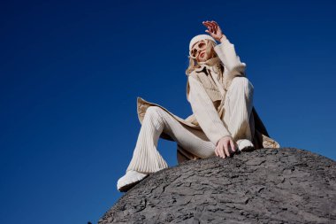 Kadın, taşın üstünde oturuyor. Mavi gökyüzü, şapka manzarasında.