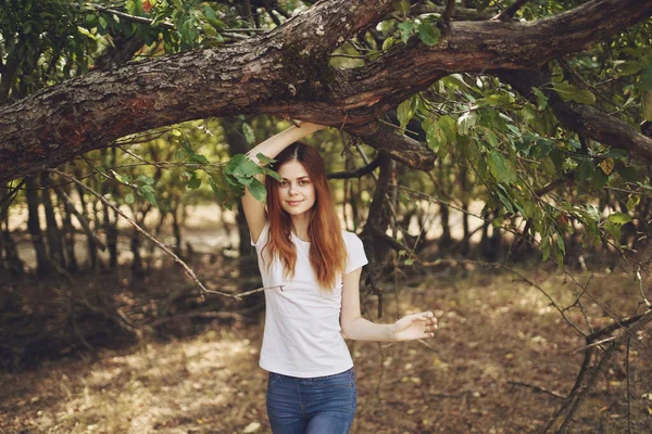 Kvinna i t-shirt och jeans står nära ett träd med gröna blad på naturen i en park — Stockfoto